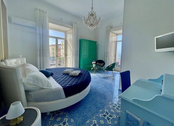 Un dormitorio con cama curvada, armario verde, televisor y vista a edificios a través de amplios ventanales.