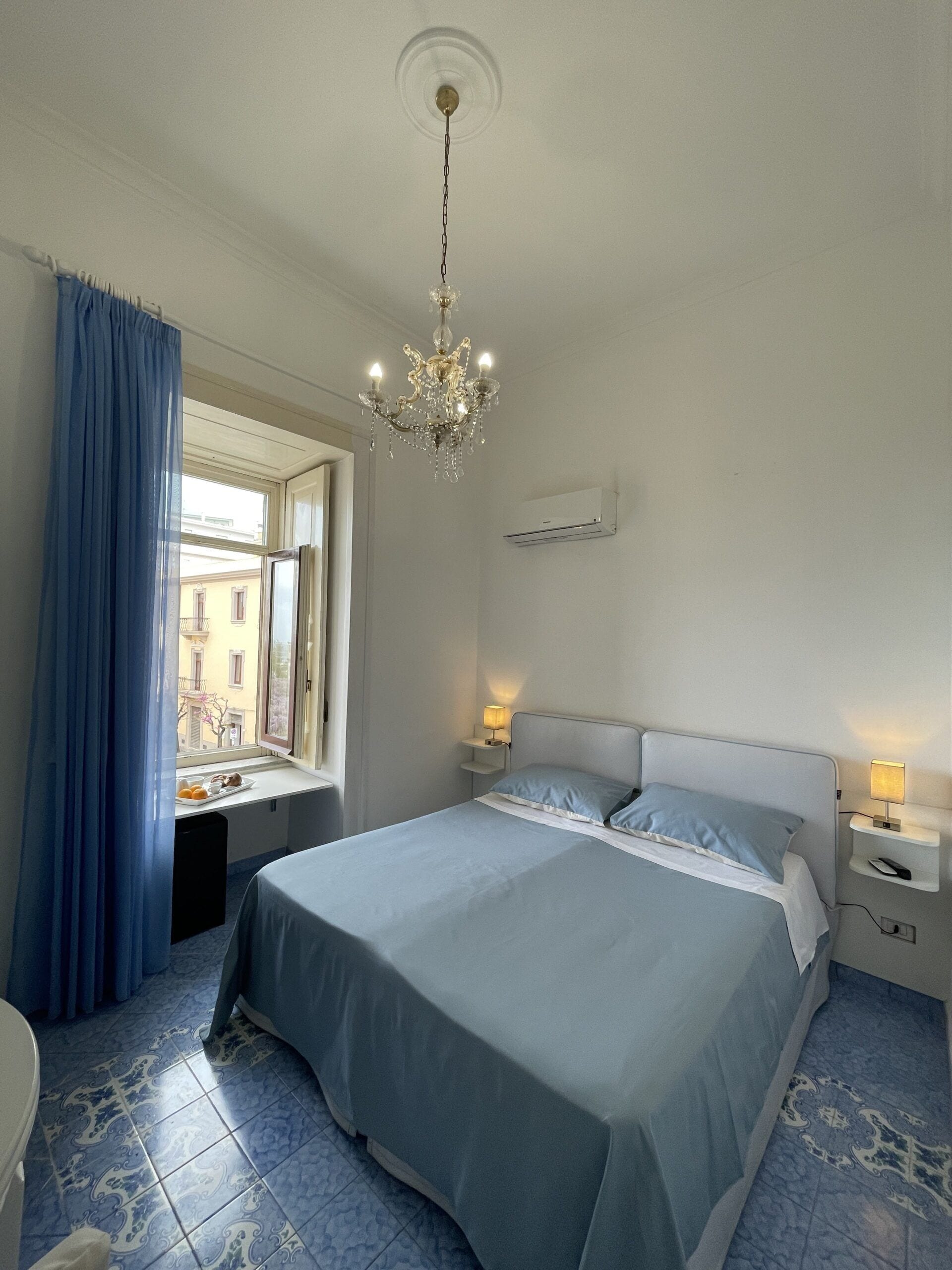 Ein Hotelzimmer mit einem großen Bett, einer Kristallleuchter, einem Fenster mit Vorhängen und einer Klimaanlage.