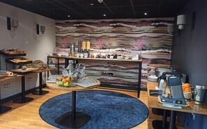 Zona de desayuno tipo buffet con una variedad de alimentos y bebidas, alfombra azul y un revestimiento de pared con textura en el fondo.