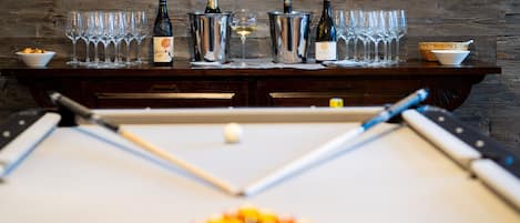 A pool table with billiard balls and cues, a set of wine bottles and glasses on a wooden table, and a bowl of snacks.