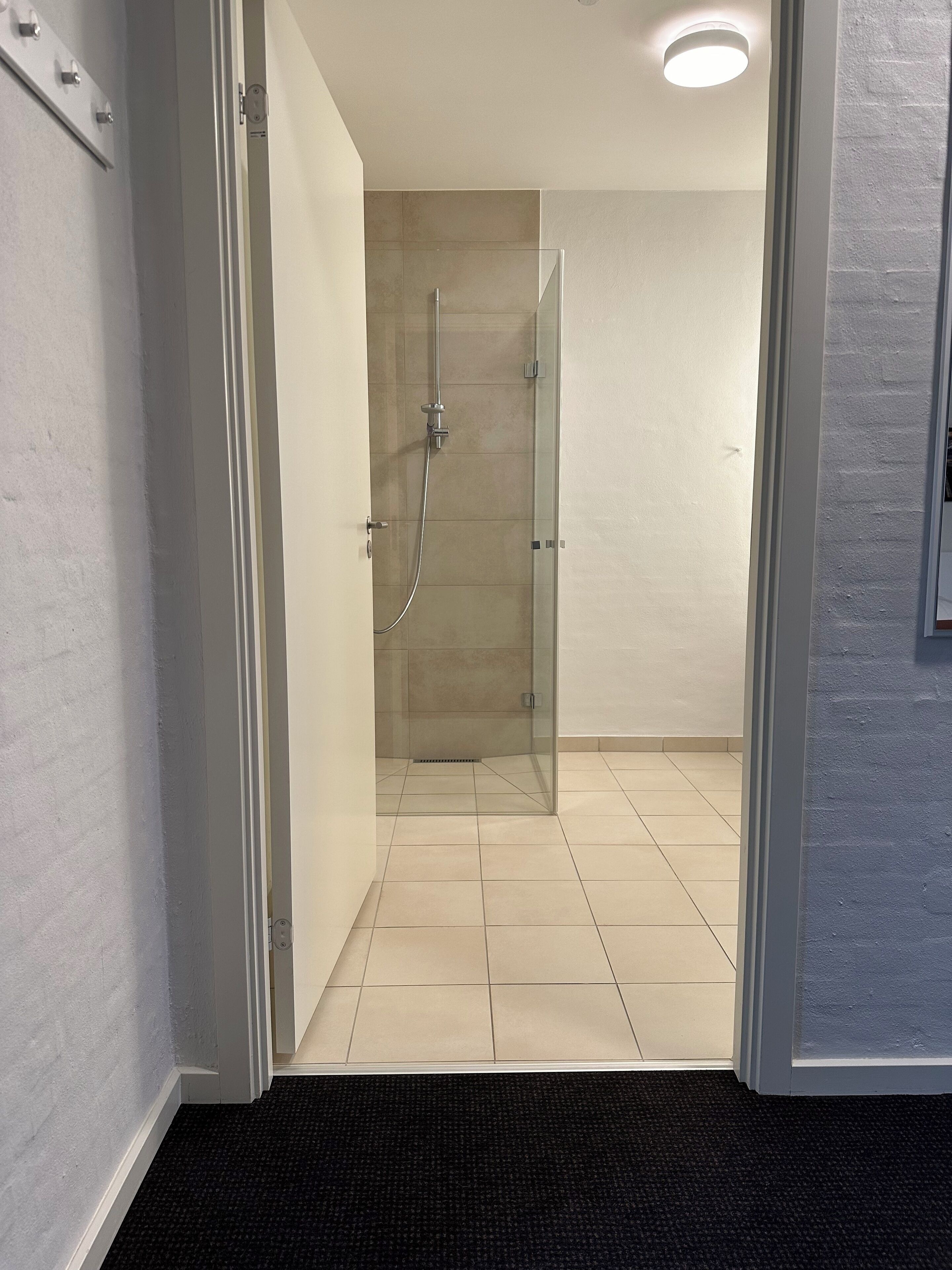 Een moderne badkamer met een douchecabine van glas, een vloer met witte tegels en een zwarte, geribbelde mat bij de ingang.