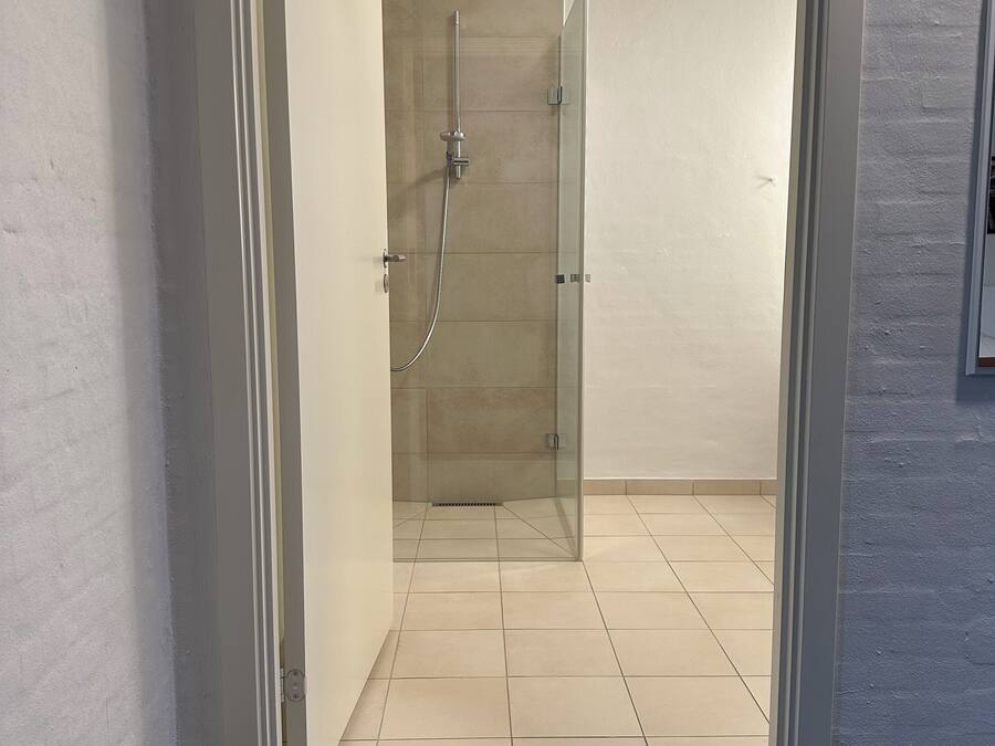 Een moderne badkamer met een douchecabine van glas, een vloer met witte tegels en een zwarte, geribbelde mat bij de ingang.