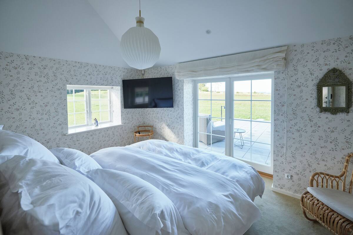 Un dormitorio con una cama grande, un televisor instalado en la pared, una ventana con vistas al exterior y una silla de mimbre.