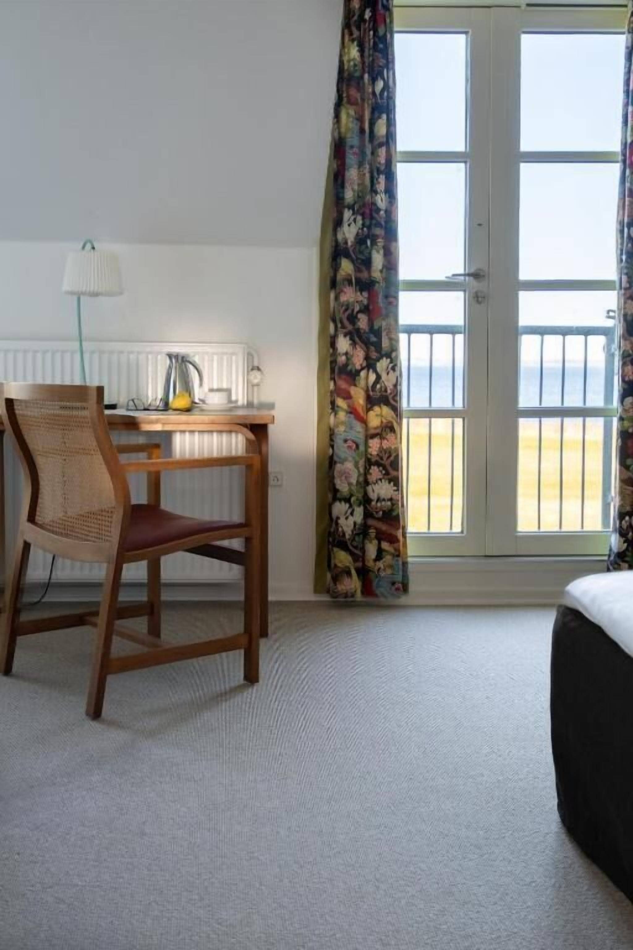 A hotel room with a wooden desk, a chair, a bed, a radiator, and a view of the beach through the window.