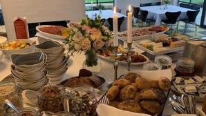 A buffet table with various food items, including fruits, cheeses, and breads, in a room with large windows and a TV in the background. - Borre Knob Fjordhotel (Horsens)