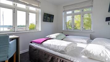 Un dormitorio con cama, escritorio y silla. Cuenta con ventanas amplias con persianas y un televisor fijado a la pared.