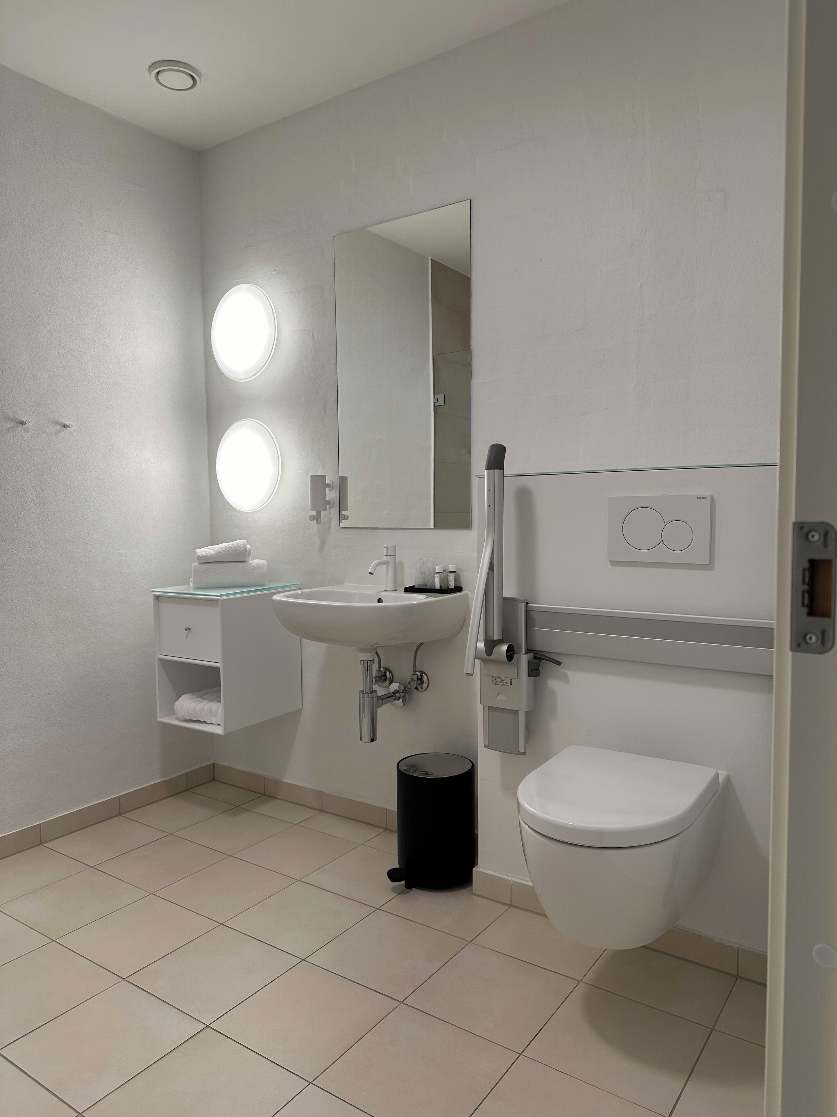 Un baño moderno con lavamanos blanco, espejo y retrete. Hay dos luces iluminadas sobre el espejo y un cubo de basura negro junto al retrete.