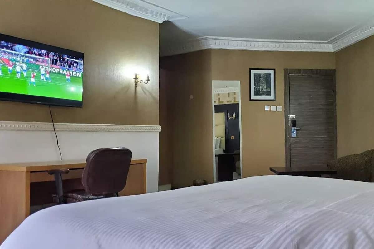 Habitación de hotel con una cama grande, un escritorio con una silla, un televisor mostrando un evento deportivo y una puerta con un espejo y cuadros.