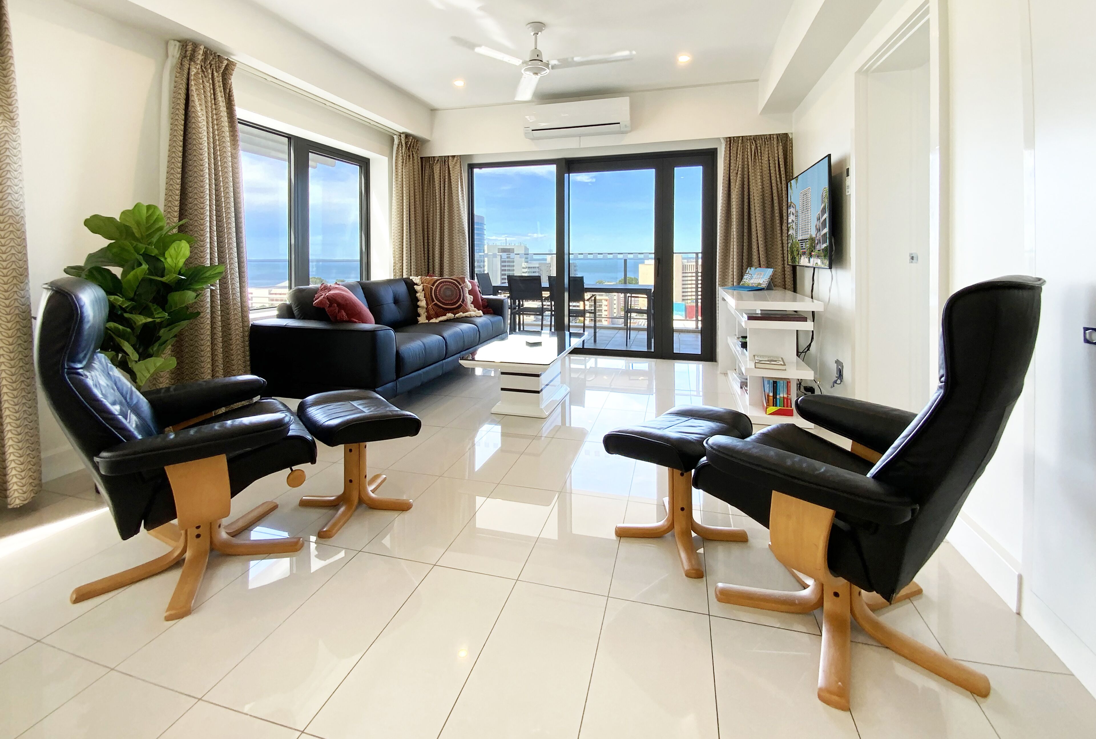 A modern living room with a black leather sofa, two black leather chairs, a coffee table, and a large window with a view of the city.