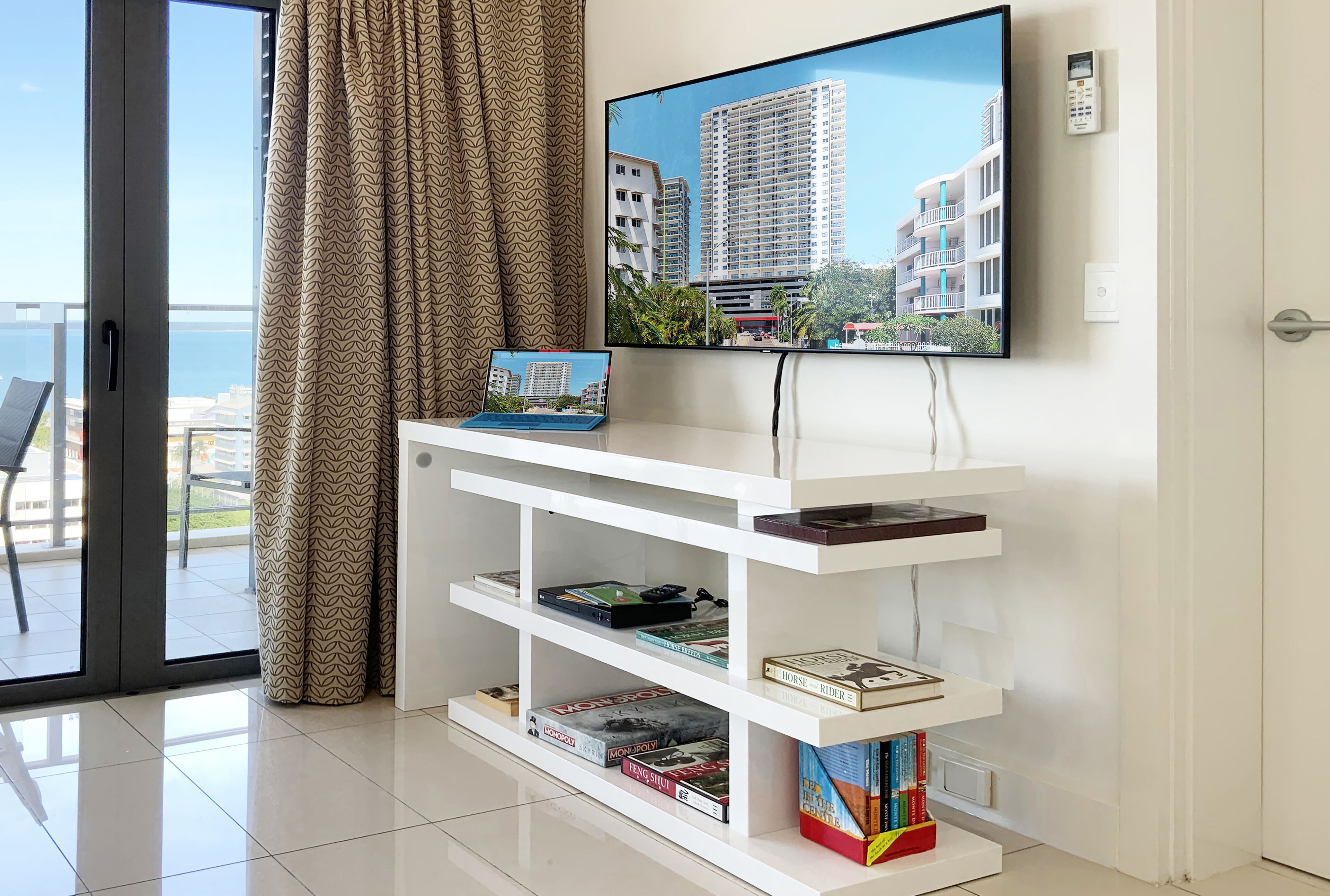 Una sala moderna con un televisor de pantalla plana montado en la pared, un mueble blanco con libros y vistas a un paisaje urbano a través de un ventanal grande.