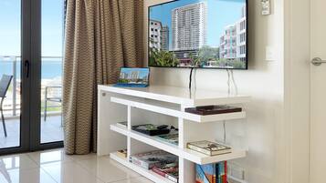 Una sala moderna con un televisor de pantalla plana montado en la pared, un mueble blanco con libros y vistas a un paisaje urbano a través de un ventanal grande.