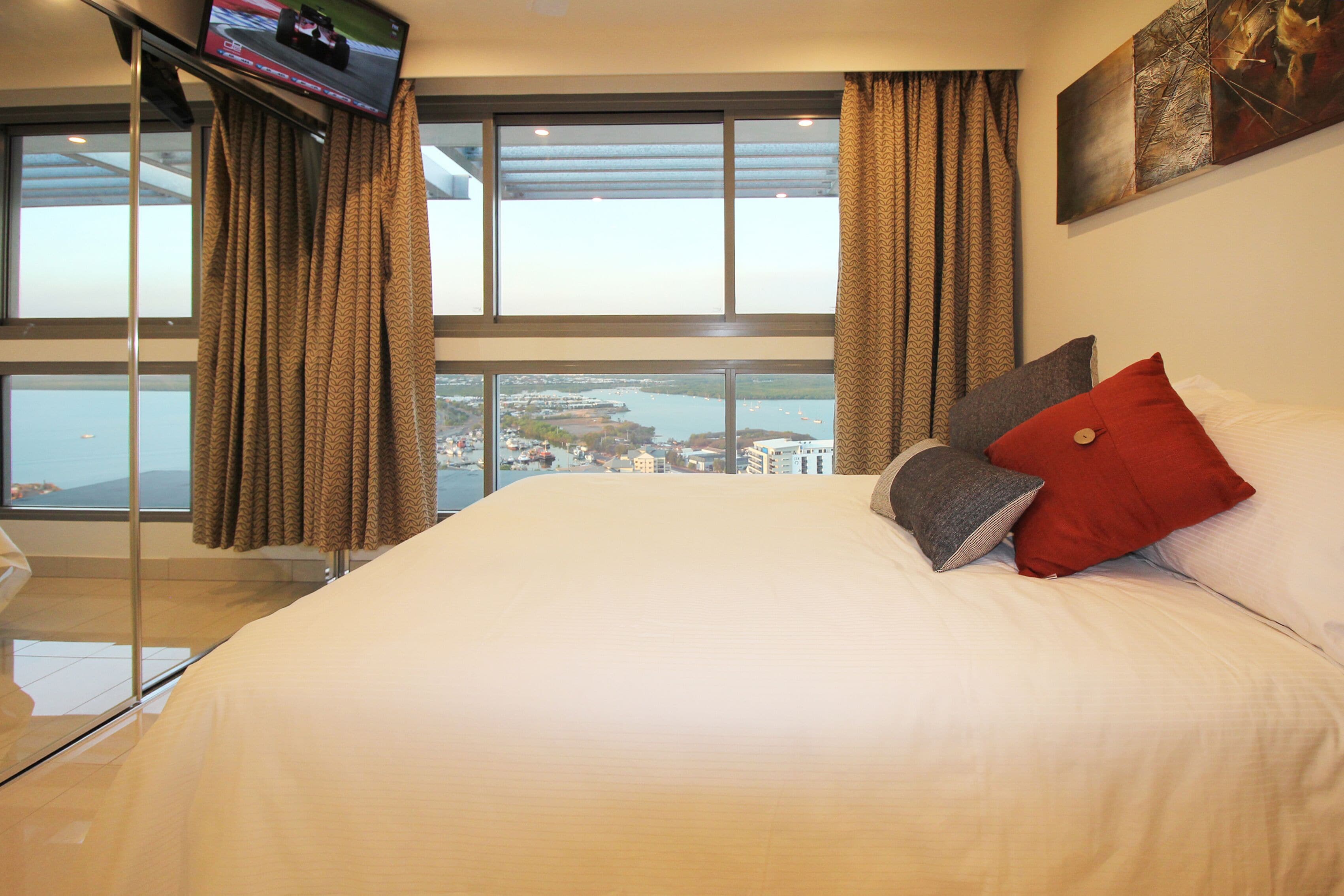 A hotel room with a large bed, a TV mounted on the wall, and a view of a cityscape through large windows.