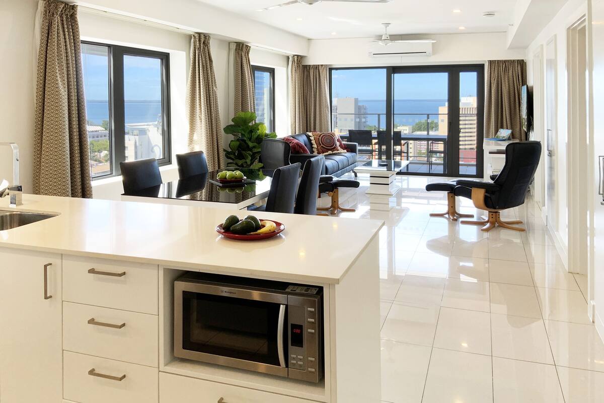 A modern kitchen with a built-in microwave, a dining area with a table and chairs, and a large window offering a view of the sea.