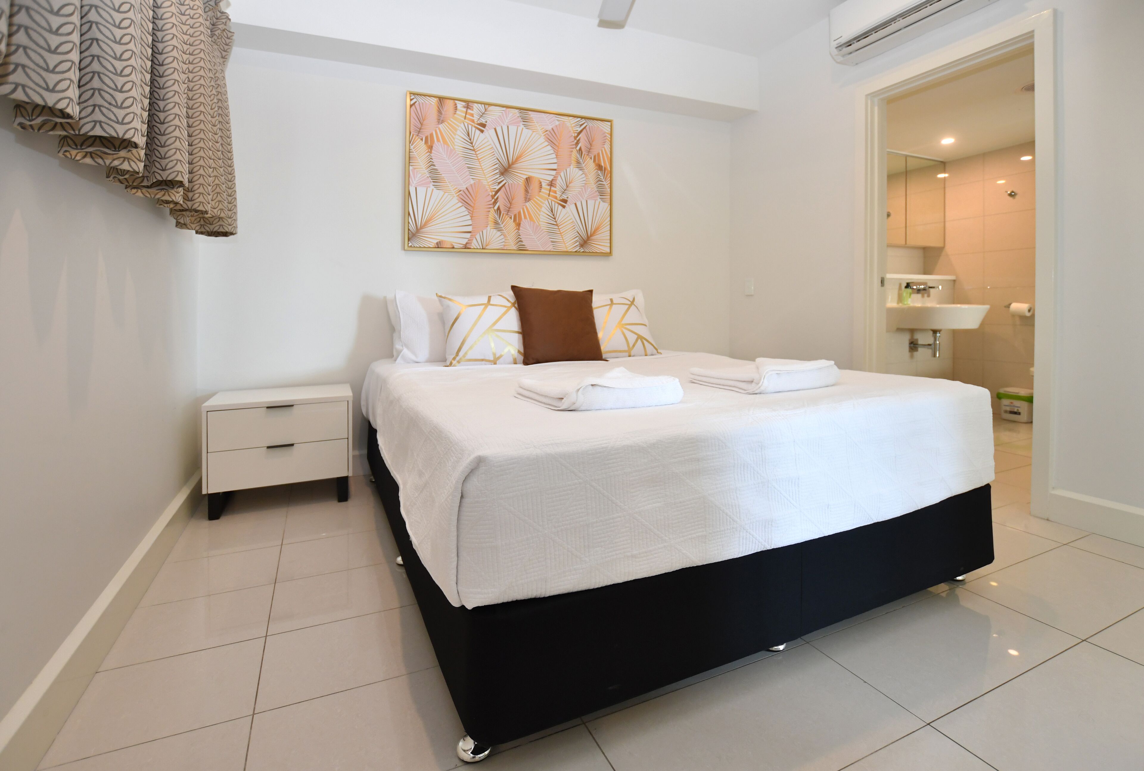 A modern bedroom with a large bed, a nightstand, a wall-mounted artwork, and a bathroom visible through an open door.