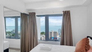 A hotel room with a large bed, two windows with curtains, a view of the city and sea, and a mirror on one wall. - Penthouse Pads (Darwin)