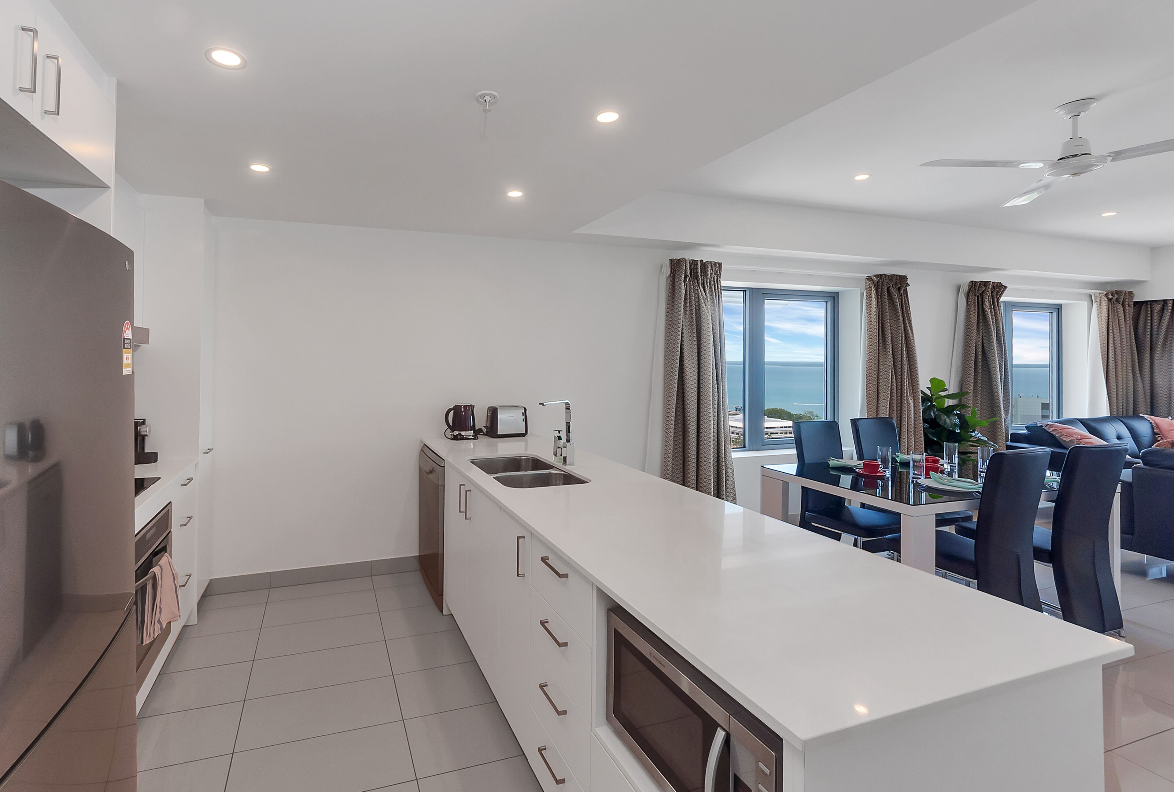 A modern kitchen with a central island, a dining area with a table and chairs, and a living area with a sofa and curtains.