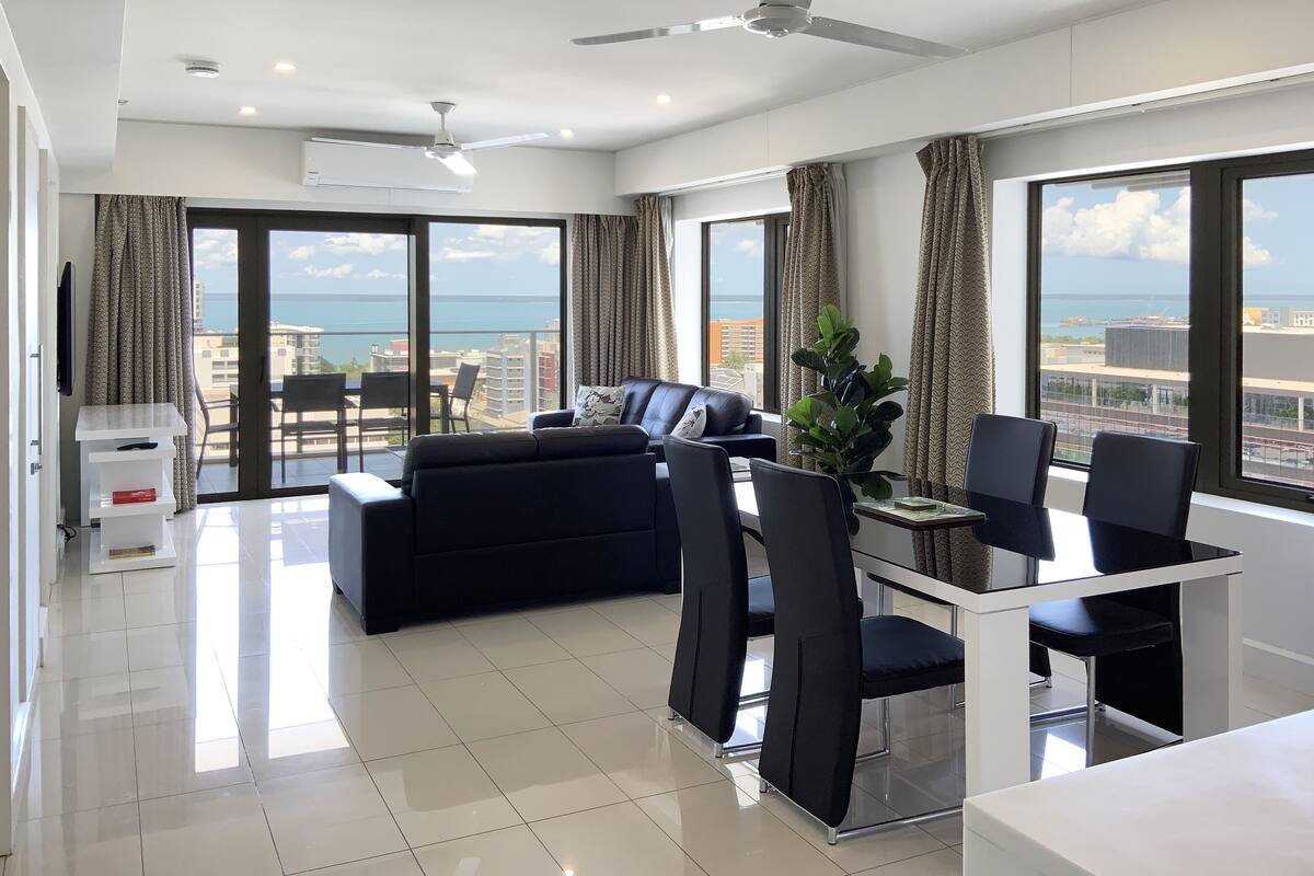 A modern living room with a black sofa, a glass dining table, and chairs. There is a ceiling fan, a TV, and a large window with a view of the sea.