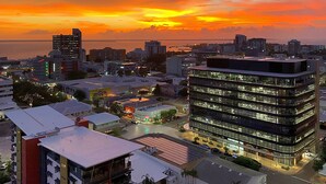 A cityscape at sunset with modern buildings, a waterfront, and a large building with multiple floors and balconies. - Penthouse Pads (Darwin)