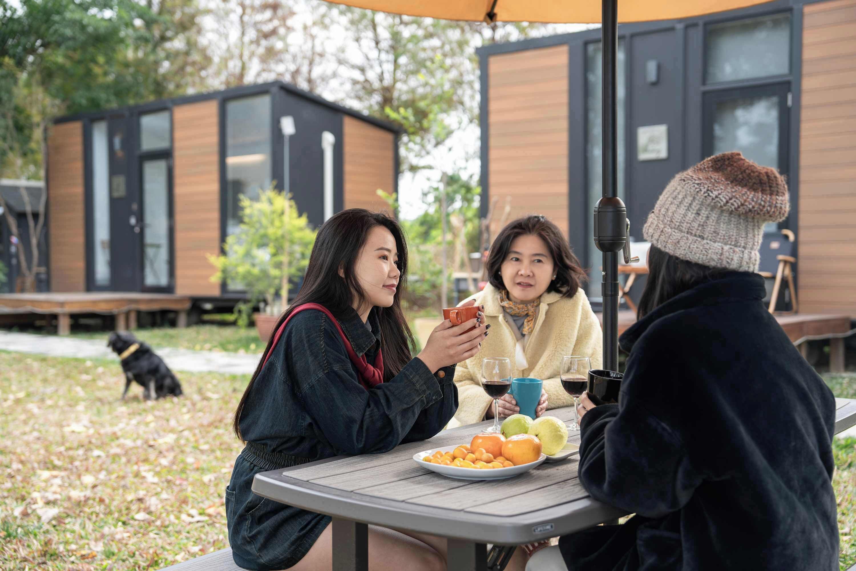 A group of people sitting at an outdoor table with a plate of fruit and drinks, in front of modern wooden buildings.