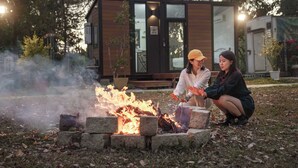 Zwei Personen sitzen am Lagerfeuer in einem Garten, im Hintergrund ein modernes Haus.