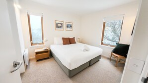 A modern hotel room with a large bed, two bedside tables, a chair, and two windows with views of trees.