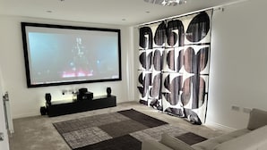 A modern living room with a large flat-screen TV mounted on the wall, a black entertainment unit below, a white sofa, and a patterned curtain. - Immaculate 4-bed House in Horley (Horley)