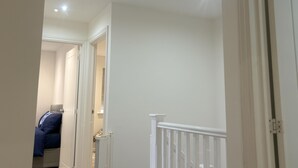 A hallway with white walls and a white door frame. There is a radiator on the wall and a bed with blue bedding in the background. - Immaculate 4-bed House in Horley (Horley)