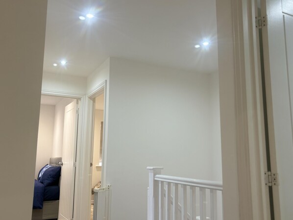 A hallway with white walls and a white door frame. There is a radiator on the wall and a bed with blue bedding in the background. - Immaculate 4-bed House in Horley (Horley)