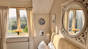 A bedroom with a bed, a window with curtains, a mirror, and a vase with flowers. - Charming 3 Bedroom Family Cottage - Dogs Welcome (Beaumaris)