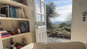 A cozy living room with a beige sofa, a bookshelf filled with books, and a view of a garden through an open door. - Charming 3 Bedroom Family Cottage - Dogs Welcome (Beaumaris)
