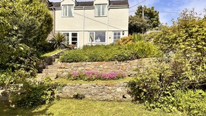 A two-story house with a stone staircase leading to the front door, surrounded by lush greenery and flowering plants. - Charming 3 Bedroom Family Cottage - Dogs Welcome (Beaumaris)