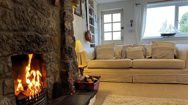 A cozy living room with a stone fireplace, a beige sofa, and a view of the outdoors through a window. - Charming 3 Bedroom Family Cottage - Dogs Welcome (Beaumaris)
