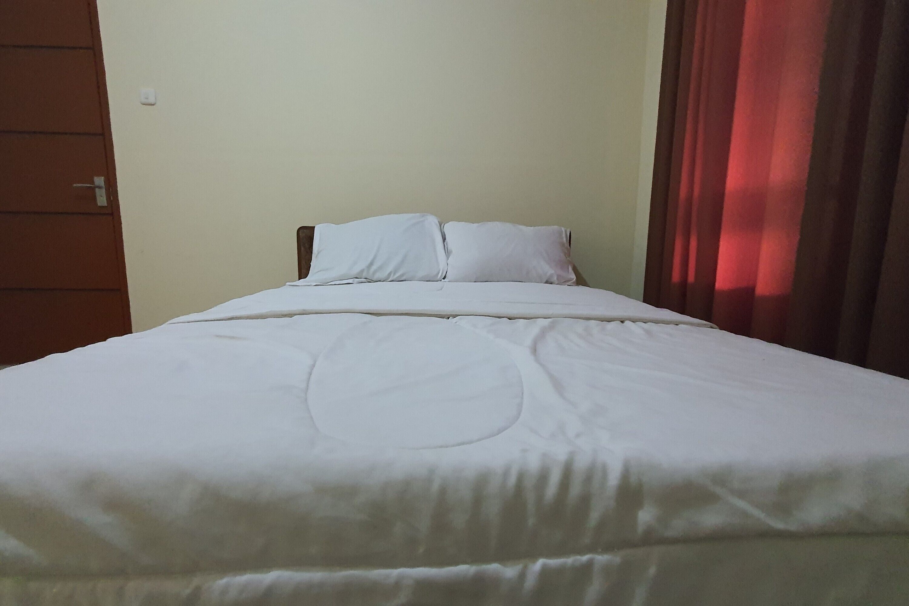 A neatly made bed with a white comforter and pillows, a wooden headboard, and a red curtain partially drawn to the side.