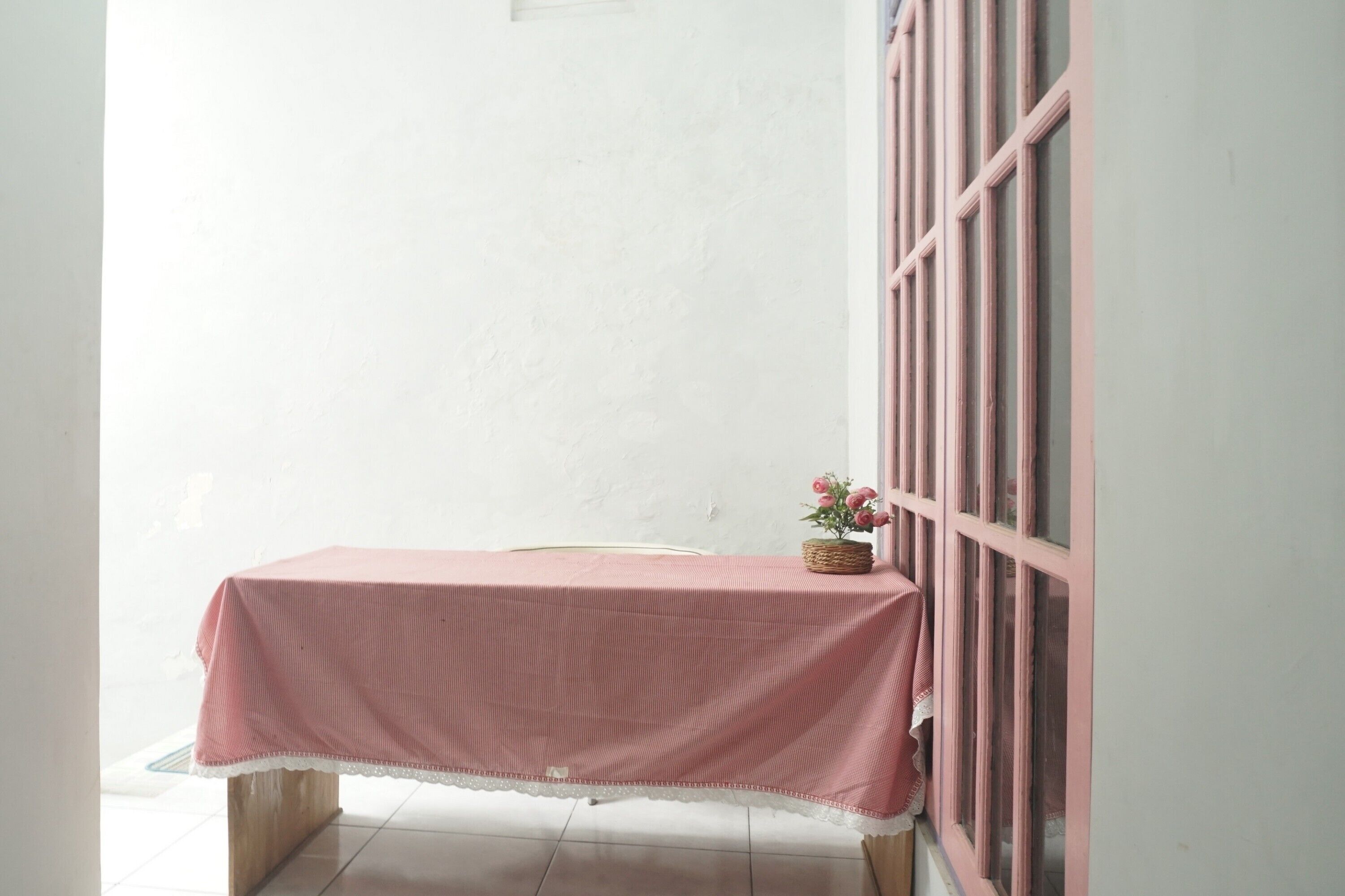 Uma pequena mesa com toalha rosa e um cesto de flores em frente a uma parede branca.