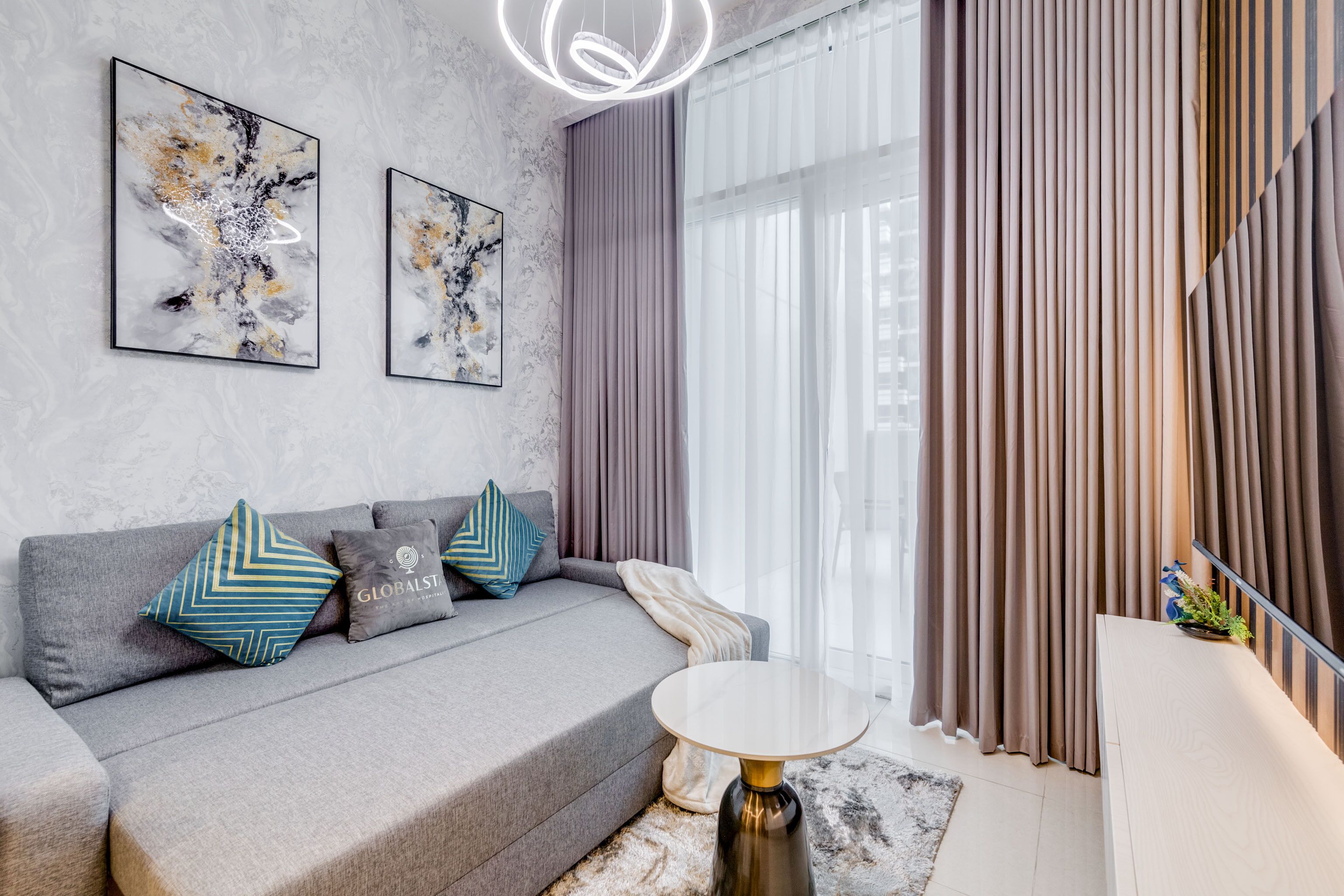 A modern living room with a grey sofa, decorative pillows, abstract wall art, a round coffee table, and sheer curtains.