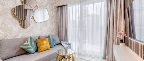 A modern living room with a grey sofa, decorative pillows, a round glass coffee table, and a large window with sheer curtains.