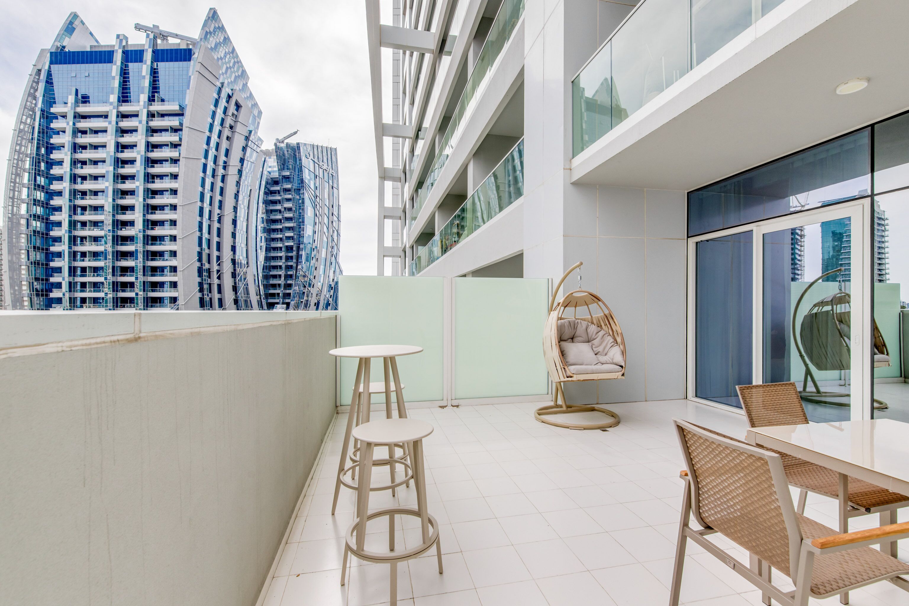 A modern outdoor balcony with a white tiled floor, wicker furniture, and a view of high-rise buildings.