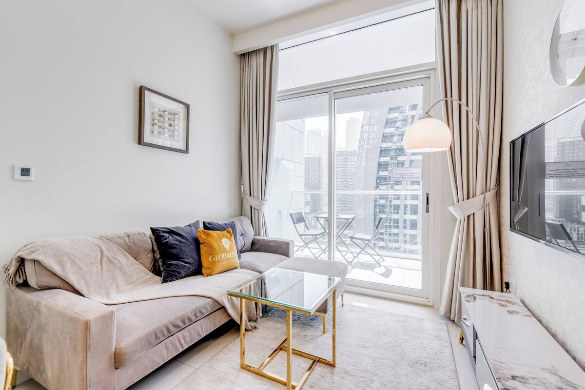 A modern living room with a glass coffee table, a sofa with cushions, a TV, and a large window with curtains.