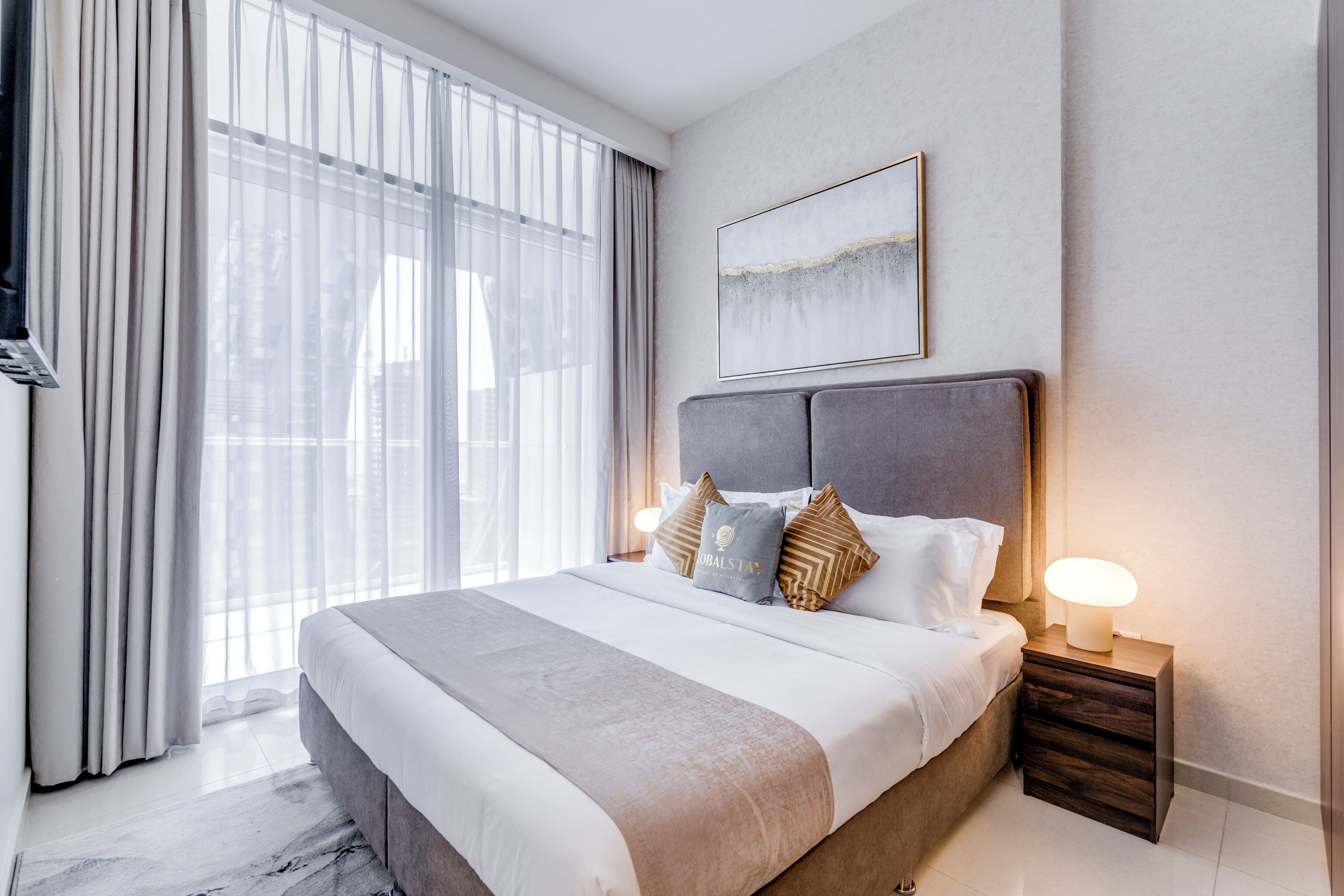 A modern hotel room with a large bed, a nightstand with a lamp, and a view of the city through large windows.