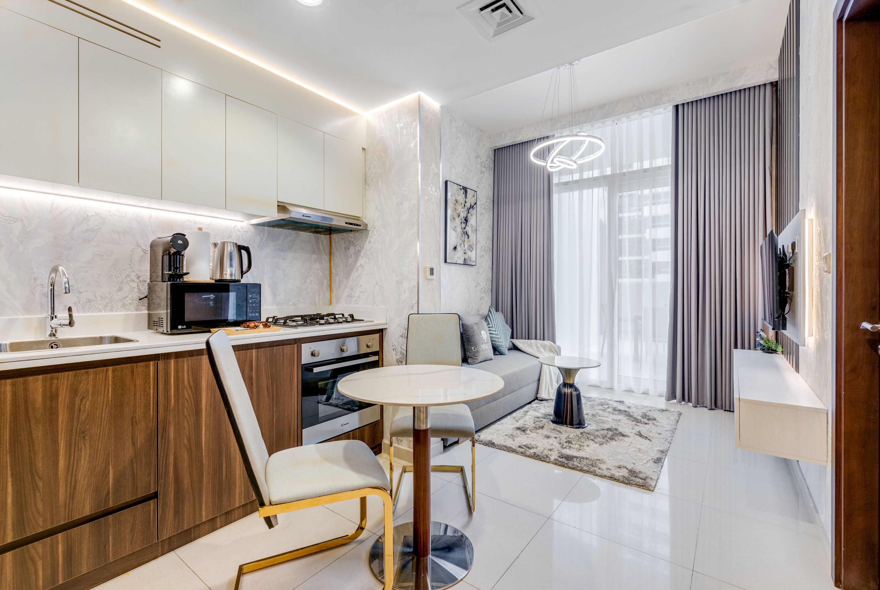 A modern kitchen with a built-in stove, microwave, and coffee maker. There is a dining area with a round table and two chairs. The room has a large window with curtains and a ceiling light fixture.