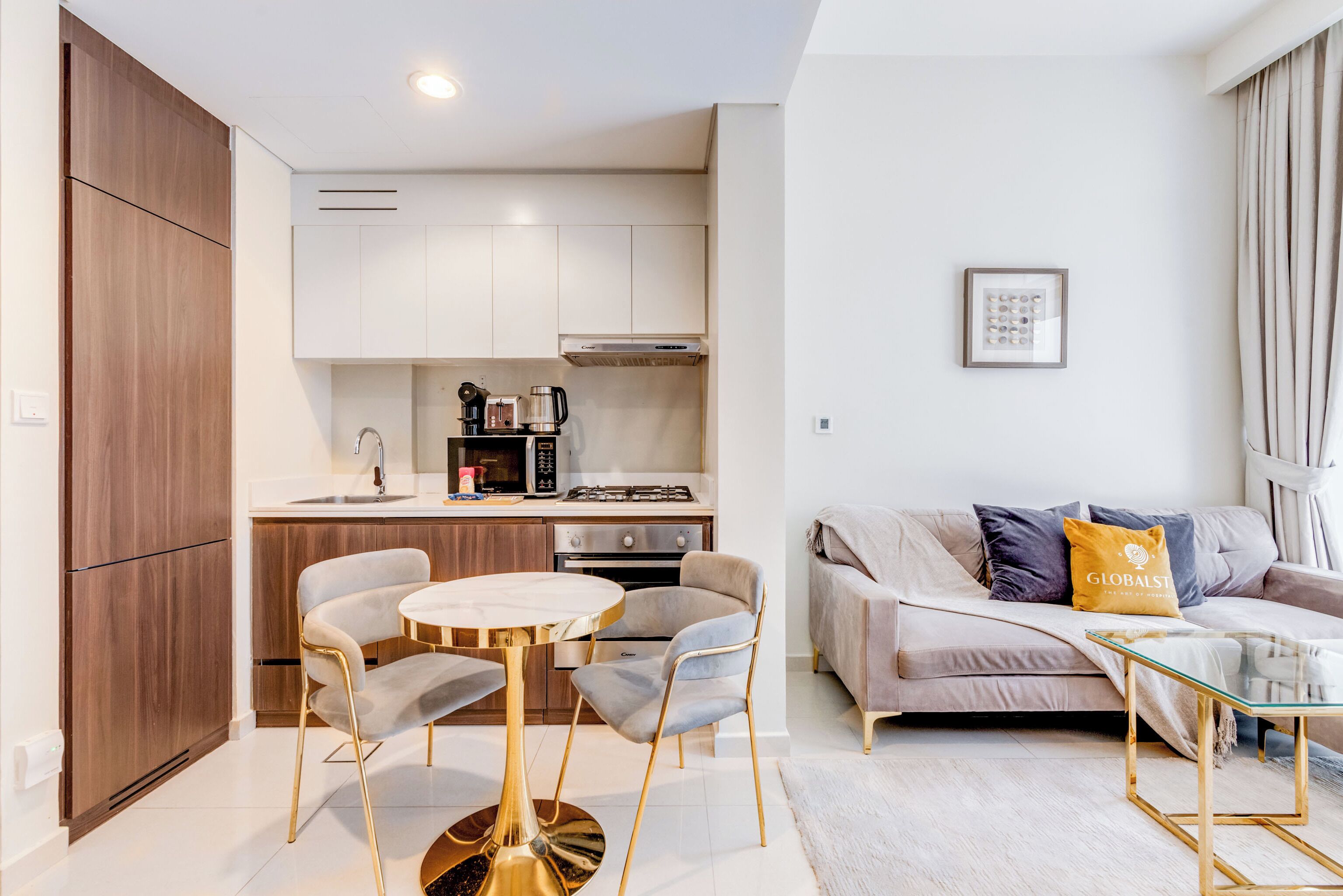 A modern kitchen with white cabinets, a gas stove, and a coffee maker. There is a dining area with a round table and chairs, and a living area with a sofa and a glass coffee table.