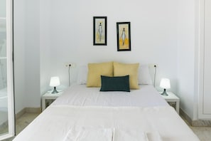 Un dormitorio con una cama, dos mesitas de noche con lámparas, un ventanal y dos cuadros en la pared.