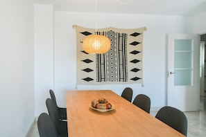 A dining area with a wooden table, black chairs, a woven hanging light fixture, and a patterned tapestry on the wall.