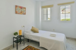 A simple, clean room with a single bed, a small table with a plant, a colorful map on the wall, and a window with a view of a balcony.