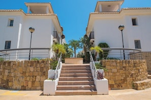 A set of outdoor stairs leading to a white building with balconies, surrounded by stone walls and palm trees.