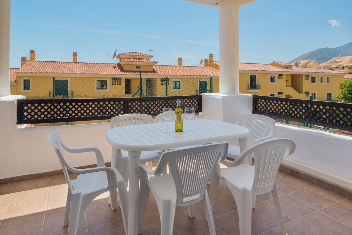 A covered outdoor patio with a round table and white plastic chairs, overlooking a row of yellow buildings with red roofs.