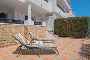 A modern building with a stone wall, a hedge, and a tiled patio with lounge chairs.