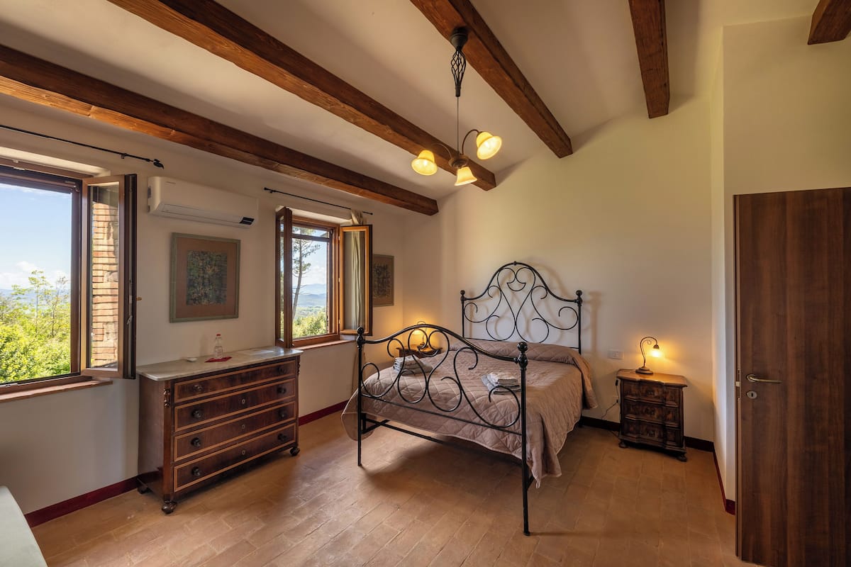 Een slaapkamer met een houten plafond, een bed met metalen frame, een dressoir, een nachtkastje en een raam met uitzicht.
