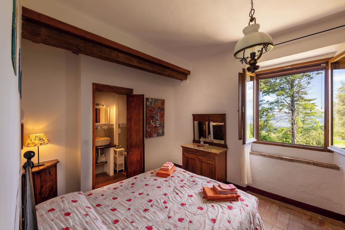 Een slaapkamer met een bed, een dressoir, een spiegel en een raam met uitzicht op bomen.