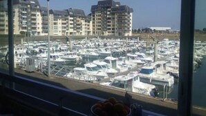 Un port de plaisance où sont amarrés de nombreux bateaux, avec en arrière-plan des immeubles d’habitation, et au premier plan, une corbeille remplie d’oranges posée sur une table.